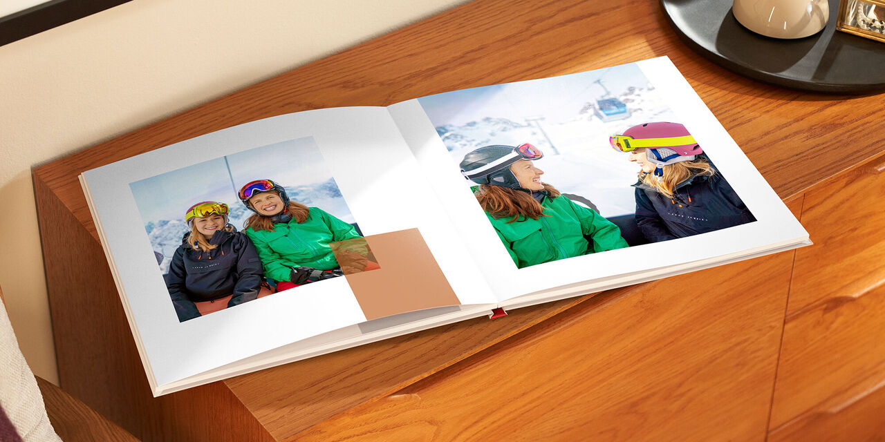 Auf einem Holzschrank liegt das aufgeklappte Fotobuch. Beide Seiten zeigen Fotos von zwei Frauen auf der Skipiste.
