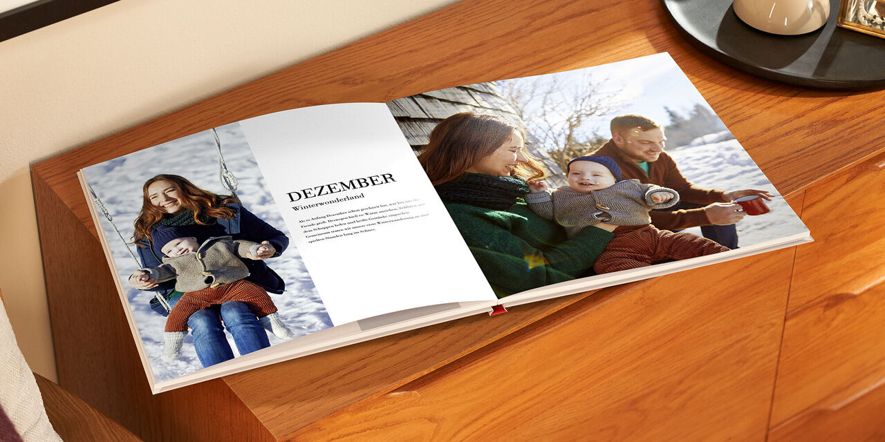 Auf einem Holzschrank liegt das aufgeklappte Fotobuch. Auf der linken Seite befinden sich ein Bild von einer Frau und einem Kind und ein Textblock mit der Überschrift „Dezember“. Auf der rechten Seite ist die Familie auf einem vollflächigen Foto zu sehen.