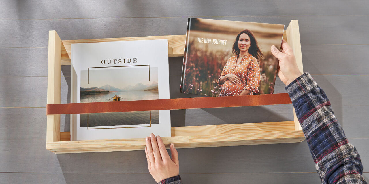 Eine Person legt ein Fotobuch mit dem Titel "The New Journey" und eine weitere Seite mit dem Wort "Outside" in eine Holzbox. Das Fotobuch hat ein schönes Cover mit einer liebevollen, besonderen Gestaltung.