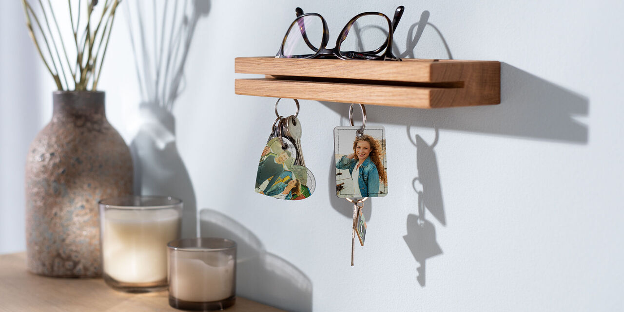 Die Schlüsselanhänger mit Foto in Herz- und Rechteck-Form hängen an einem Schlüsselbrett. Auf dem Schlüsselbrett liegt eine Brille mit schwarzem Rahmen. Im Hintergrund stehen eine Vase mit Trockenblumen und zwei Kerzen.